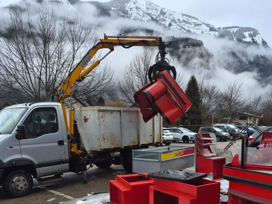 Le camion-benne de Perreau récupération, grue dépliée, charge du mobilier métallique rouge au grappin sur un parking enneigé, montagnes au fond.